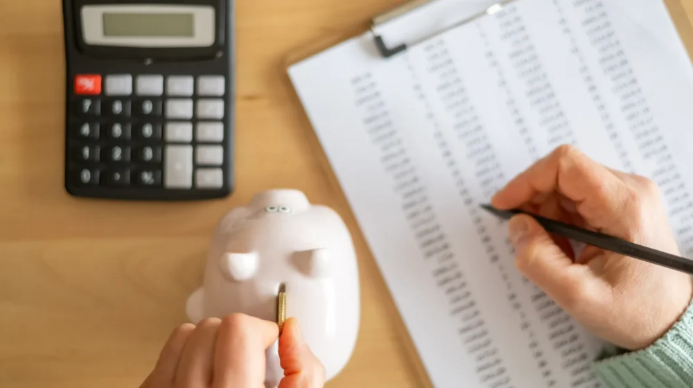 Accountant inserting a coin into piggy bank and calculating expenses with calculator and spreadsheet on a wooden desk, managing home budget and savings