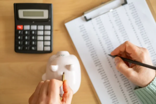 Accountant inserting a coin into piggy bank and calculating expenses with calculator and spreadsheet on a wooden desk, managing home budget and savings
